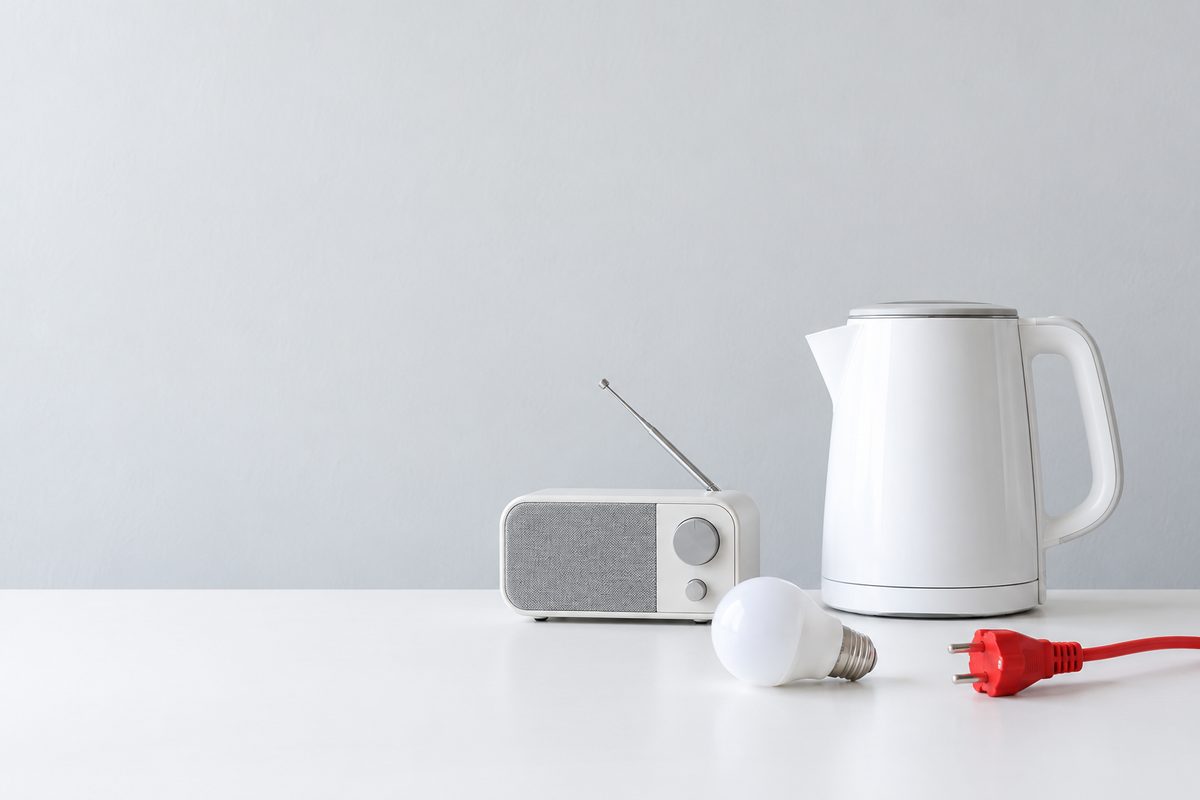 白いテーブルに並ぶ電気ケトル、ラジオ、電球と赤い電源プラグ